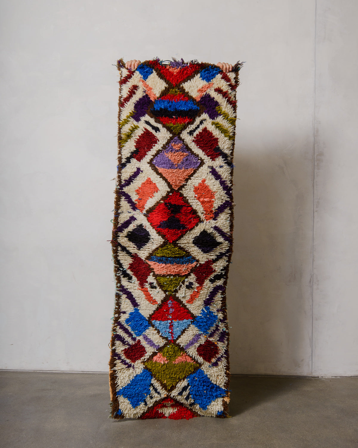Colourful boucherouite rug being held up in front of a plain background.
